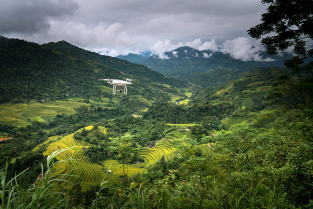 A white drone hovering over a lush green valley with terraced farmland, capturing agricultural data for precision farming. 